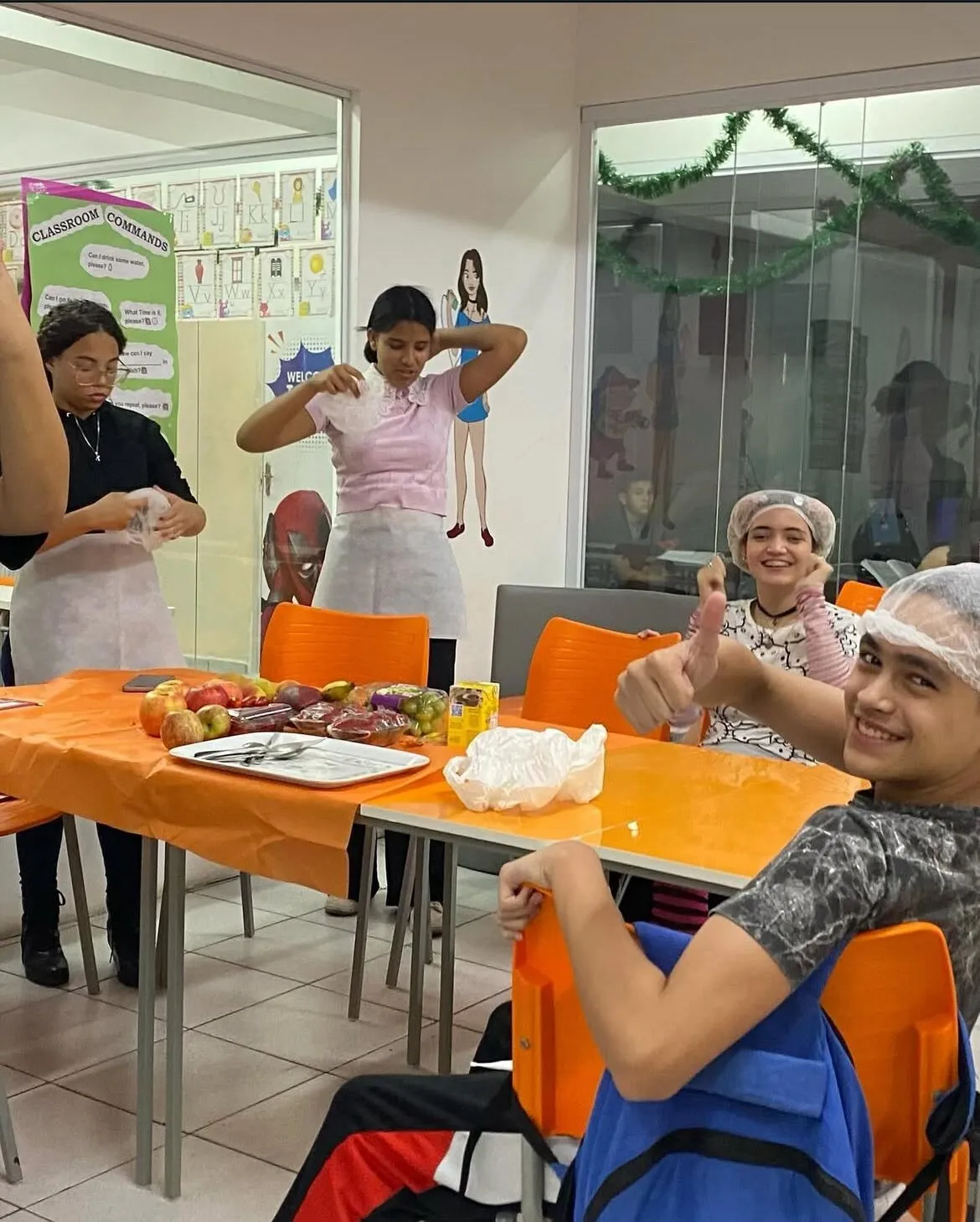 Alunos de touca preparando frutas em aula prática, um deles faz sinal de positivo